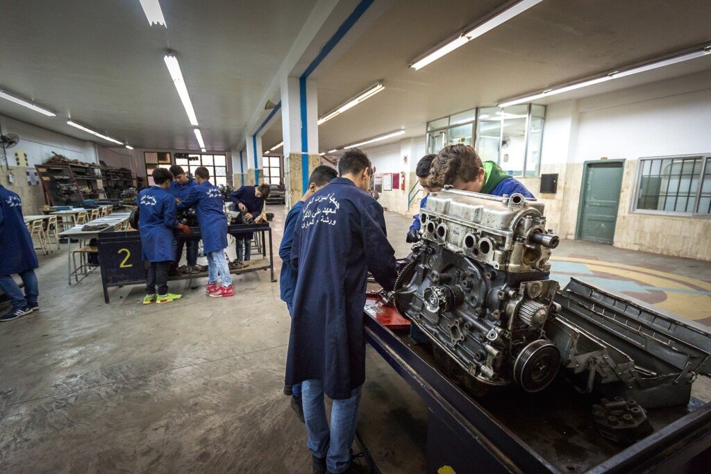 طلاب بمعاطف زرقاء يتدربون على صيانة محرك سيارة في ورشة ميكانيك المبرات المهنية | Students in blue coats training on car-engine maintenance in an Al-Mabarrat vocational mechanics workshop