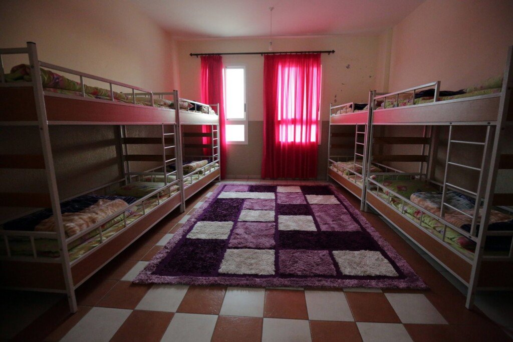 غرفة تحتوي أسرّة طابقية ونوافذ بستائر حمراء وسجاد في المنتصف | Room featuring bunk beds, red curtains, and a central carpet