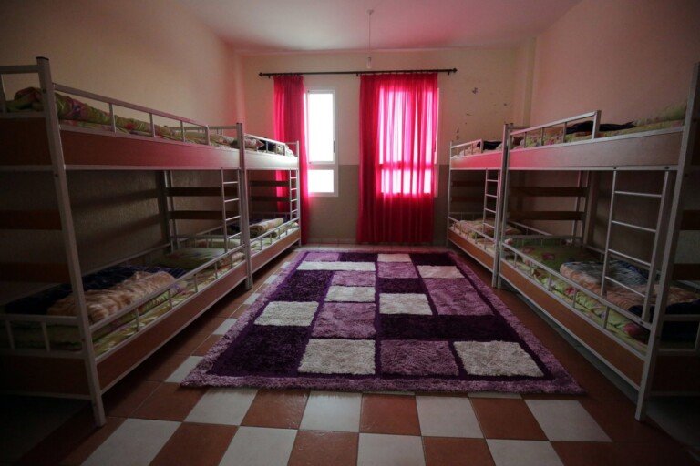 غرفة تحتوي أسرّة طابقية ونوافذ بستائر حمراء وسجاد في المنتصف | Room featuring bunk beds, red curtains, and a central carpet