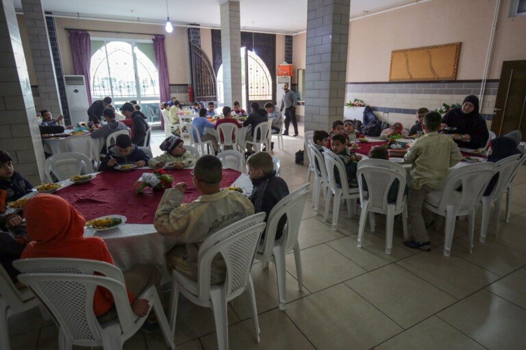 أطفال يجلسون على طاولات ويتناولون الطعام في قاعة طعام ضمن مبرة الإمام علي (ع) | Children sitting at tables having a meal in a dining hall at Imam Ali Institution