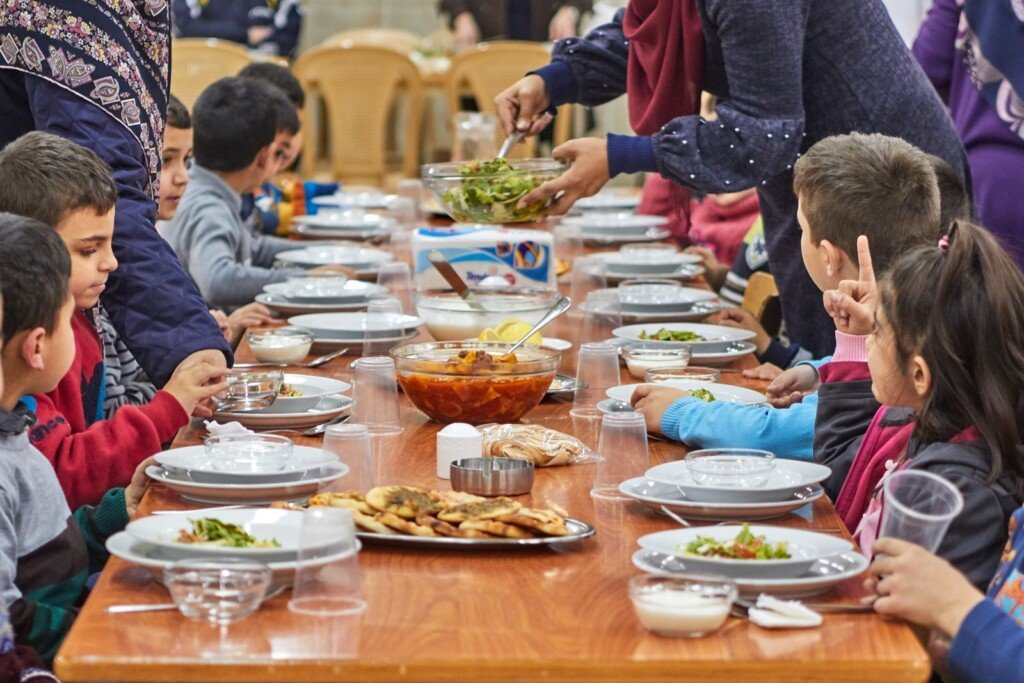 أطفال يجلسون حول طاولة ويتناولون وجبة طعام مع تقديم السلطات ضمن مبرة النبي إبراهيم (ع) في لبنان | Children sitting around a table sharing a meal with salad being served at Prophet Ibrahim Institution (AS) in Lebanon