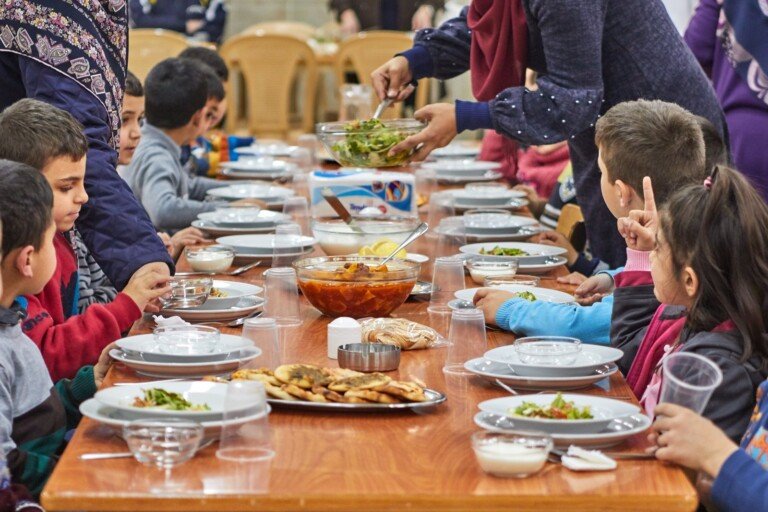 أطفال يجلسون حول طاولة ويتناولون وجبة طعام مع تقديم السلطات ضمن مبرة النبي إبراهيم (ع) في لبنان | Children sitting around a table sharing a meal with salad being served at Prophet Ibrahim Institution (AS) in Lebanon