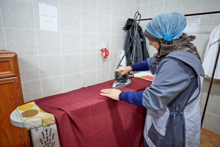 عاملة تقوم بكيّ وتجهيز الأقمشة داخل غرفة خدمات ضمن مبرة النبي إبراهيم (ع) | Staff member ironing and preparing fabrics inside a service room at Al Mabarrat Association facility