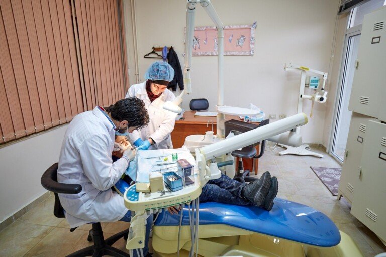 طبيب وطبيبة أسنان يقدمان العلاج لطفل داخل عيادة مجهزة ضمن برامج الرعاية الصحية التابعة لمبرة النبي إبراهيم (ع) | A dentist and assistant providing dental treatment to a child in a fully equipped clinic under Al Mabarrat Association healthcare services
