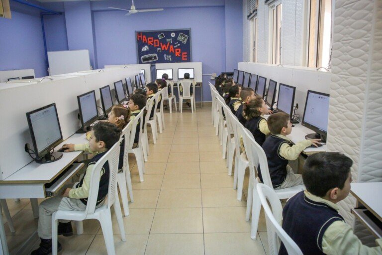 أطفال صغار يعملون على الحاسوب في مختبر مدرسة الإشراق مزين بكلمة Hardware | Young children working on computers in the lab of Al-Ishraq School decorated with the word Hardware