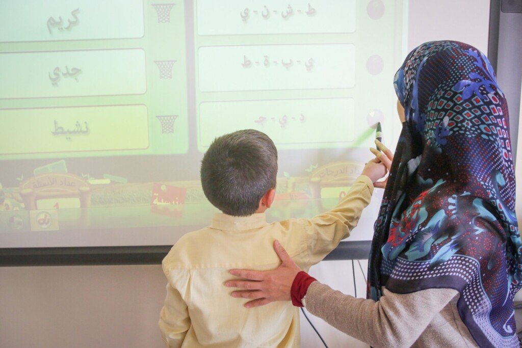 معلمة تساعد طفلاً على حل نشاط أحرف عربية على اللوح التفاعلي في مدرسة الإشراق | A teacher helping a boy solve an Arabic-letters activity on the interactive board at Al-Ishraq School
