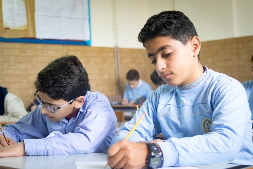طالب يجلس في صف مدرسي ويكتب على الورق أثناء الحصة الدراسية | Student sitting in a classroom writing on paper during a lesson