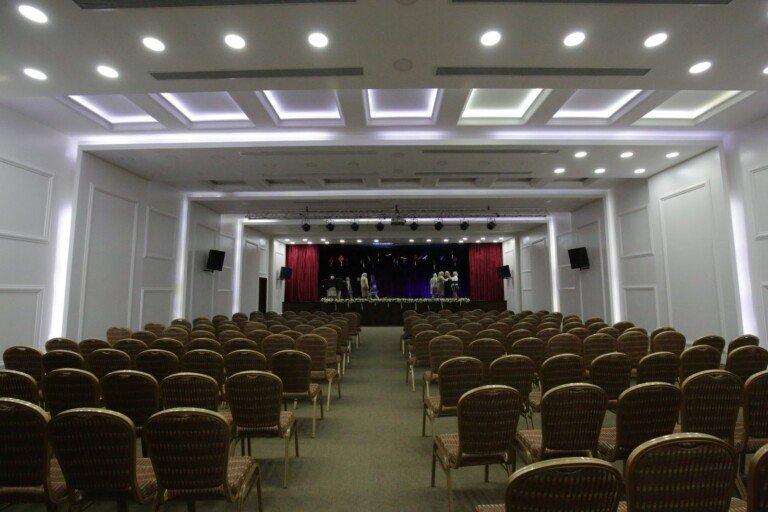 قاعة كبيرة تحتوي على مقاعد مرتبة ومسرح مجهز داخل مدرسة ثانوية الإمام علي (ع) | Large auditorium with arranged seating and a stage inside Thanaouiya Al Imam Ali (A) school