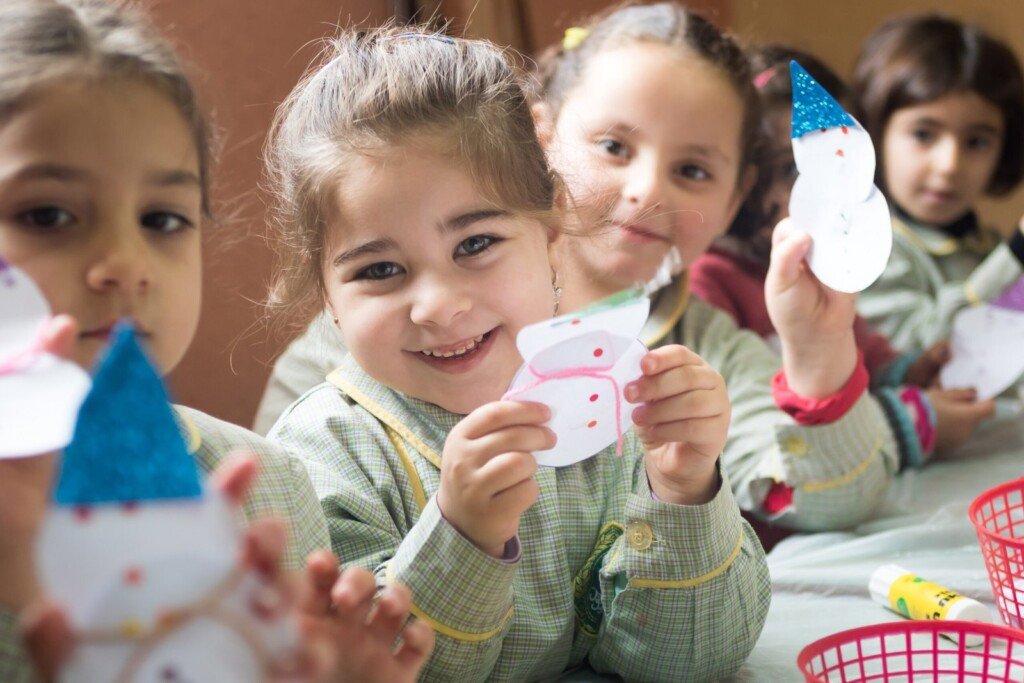 أطفال يشاركون في نشاط فني داخل صف مدرسي يحملون أعمالاً ورقية يدوية | Children participating in an art activity in a classroom holding handmade paper crafts
