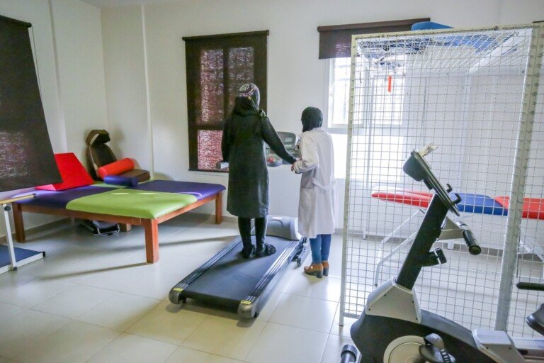 معالجة ترافق سيدة على جهاز المشي في قسم العلاج الفيزيائي بمركز المصطفى | A therapist accompanying a woman on the treadmill in the physiotherapy section of Al-Mustafa Center