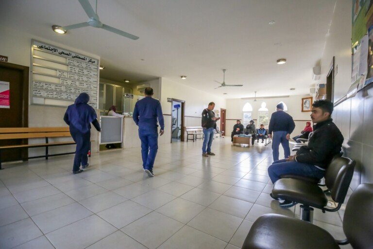 مراجعون في قاعة انتظار مركز المصطفى الصحي مع لوحة أسماء العيادات | Visitors in the waiting hall of Al-Mustafa Health Center with the clinics board