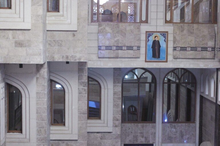 واجهة داخلية حجرية بنوافذ مقوّسة وصورة معلّقة للسيد محمد حسين فضل الله على الجدار الرخامي | A stone interior facade with arched windows and a hung portrait of Sayyed Fadlallah on the marble wall