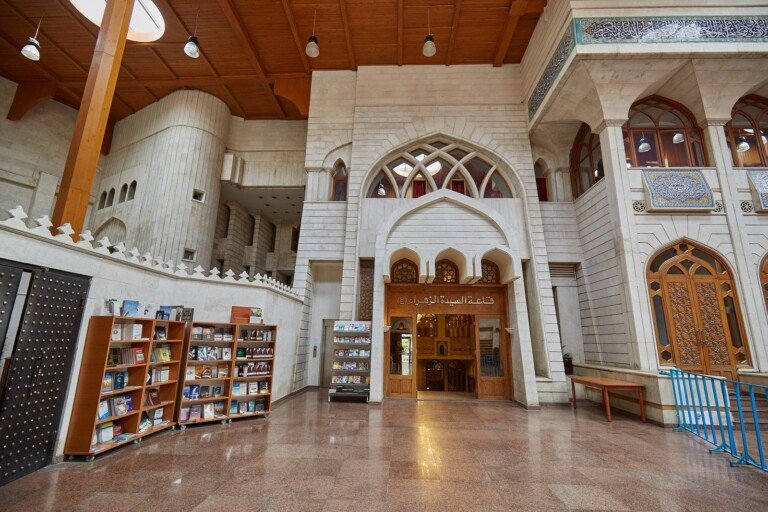 بهو رخامي واسع بأقواس وزخارف إسلامية ورفوف كتب ومدخل قاعة السيدة الزهراء | A spacious marble lobby with Islamic arches and ornamentation, bookshelves and the entrance to Sayyeda Zahraa Hall