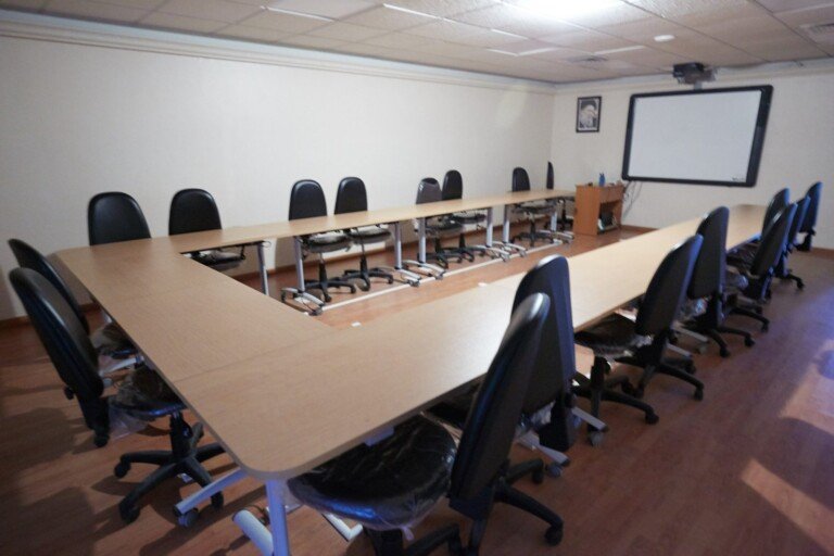 قاعة اجتماعات فيها طاولة خشبية على شكل حرف U وكراسٍ مكتبية سوداء وشاشة عرض تفاعلية | A meeting room with a U-shaped wooden table, black office chairs and an interactive whiteboard