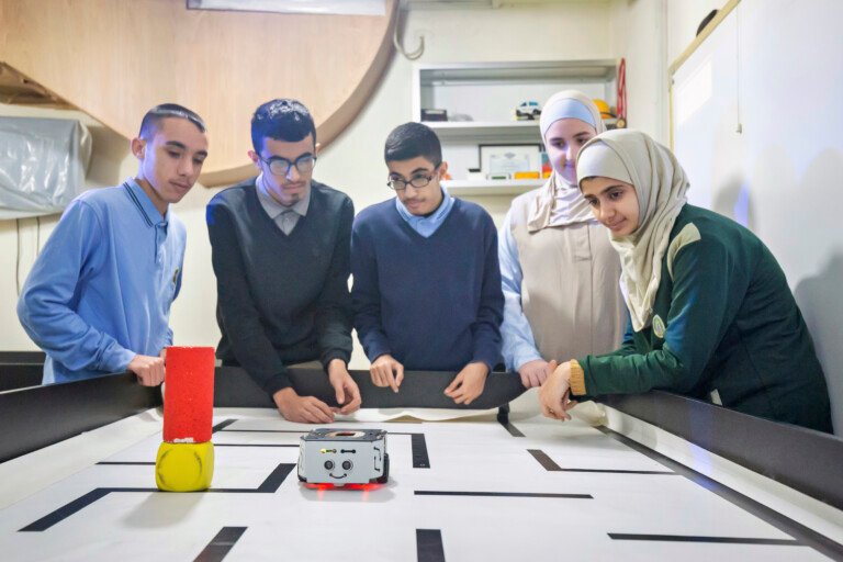 طلاب يراقبون روبوتاً صغيراً يتحرك في متاهة على طاولة في مؤسسة الإمام الهادي | Students watching a small robot navigate a maze on a table at Al-Imam Al-Hadi Institution