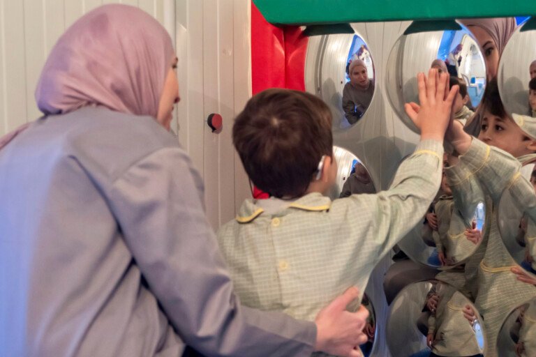 معلمة ترافق طفلاً بسماعة أذن أمام جدار المرايا المحدبة في مؤسسة الإمام الهادي | A teacher accompanying a child with a hearing aid at the convex mirror wall at Al-Imam Al-Hadi Institution