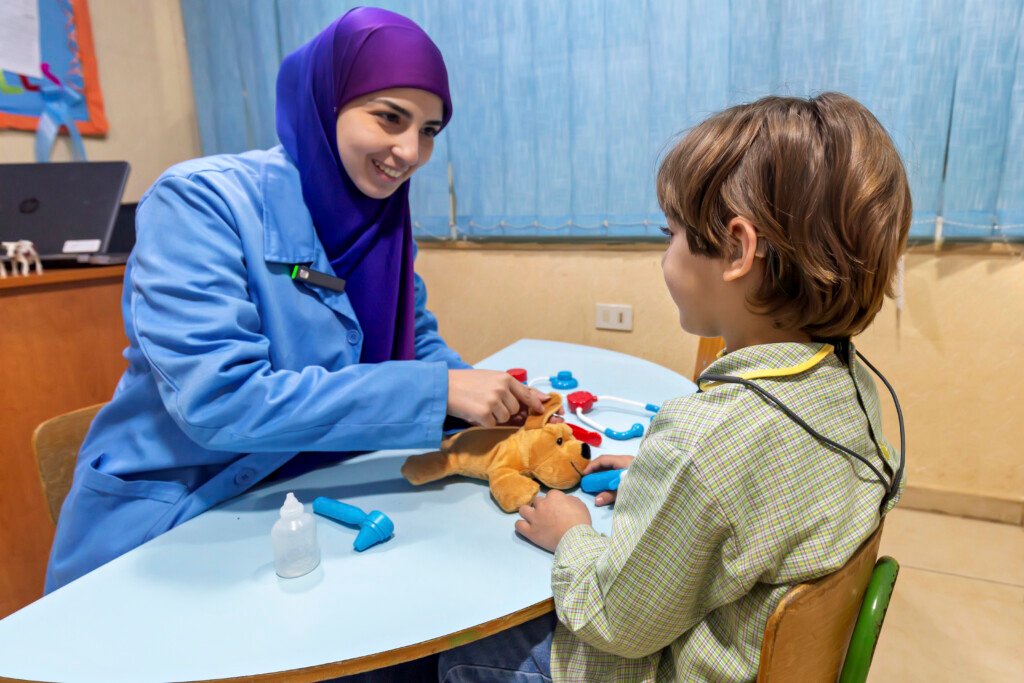 اختصاصية تجلس مع طفل بسماعة أذن وبينهما دمية ولعب طبية في مؤسسة الإمام الهادي | A specialist with a hearing-aided child, a doll and toy medical tools between them at Al-Imam Al-Hadi Institution