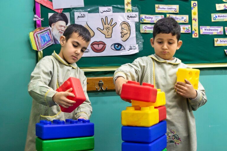 طفلان بمريول مدرسي يركبان مكعبات ملونة أمام لوحة الحواس الخمس في مؤسسة الإمام الهادي | Two children in school aprons building colored blocks before the five-senses board at Al-Imam Al-Hadi Institution