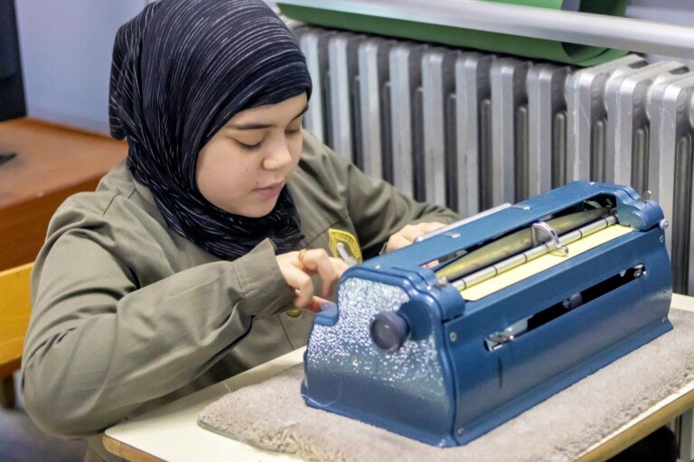 طالبة تستخدم آلة برايل زرقاء للكتابة النافرة في مؤسسات المبرات للتربية المختصة | A student using a blue Braille writer at Al-Mabarrat special-education institutions