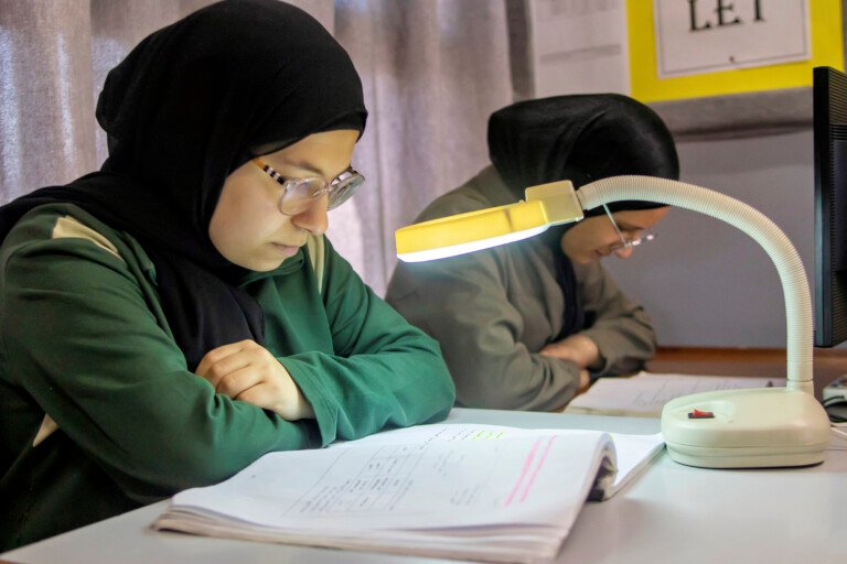 طالبتان تدرسان تحت مصباح مكتبي مكبر في مؤسسة الإمام الهادي | Two students studying under a magnifying desk lamp at Al-Imam Al-Hadi Institution