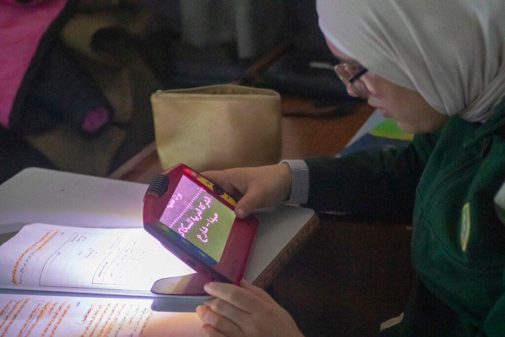 طالبة تمسك جهاز تكبير إلكترونياً يعرض نصاً عربياً مكبراً في مؤسسة الإمام الهادي | A student holding a portable electronic magnifier displaying enlarged Arabic text at Al-Imam Al-Hadi Institution