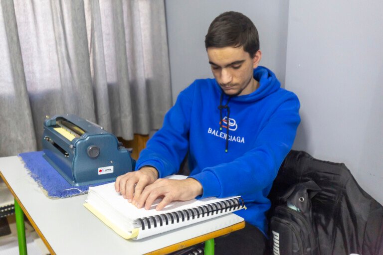 شاب يقرأ بأنامله دفتر برايل وبجانبه آلة برايل في مؤسسة الإمام الهادي | A young man reading a Braille notebook with his fingertips beside a Braille writer at Al-Imam Al-Hadi Institution