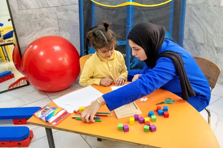 اختصاصية تدرب طفلة بمكعبات خشبية ملونة في مراكز التشخيص التربوي للمبرات | A specialist training a girl with colored wooden blocks at Al-Mabarrat educational diagnosis centers