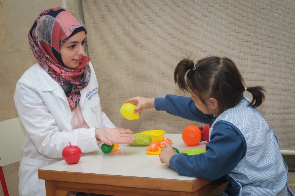 اختصاصية نطق بمعطف أبيض مع طفلة وألعاب فواكه بلاستيكية في مركز سراج | A speech therapist in a white coat with a girl and plastic fruit toys at Siraj Center