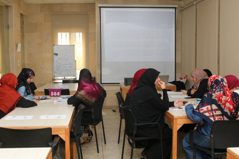 مشاركات حول طاولات في قاعة تدريب بمركز سراج أمام شاشة عرض | Participants around training tables before a projection screen at Siraj Center
