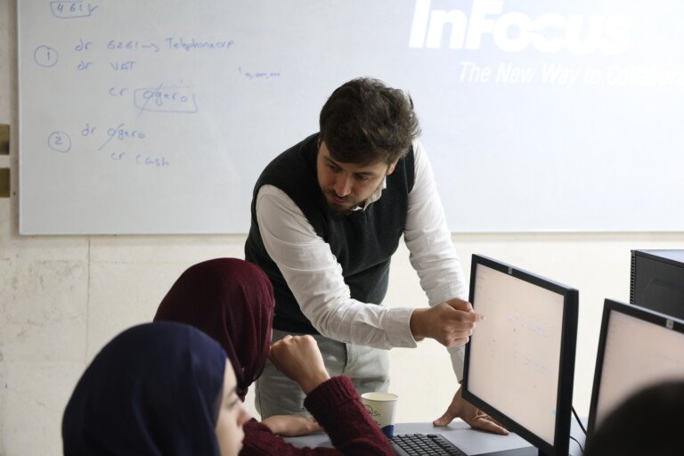 أستاذ في جامعة USAL يشرح تطبيقاً محاسبياً لطالبتين أمام الشاشة واللوح | A USAL professor explaining an accounting application to two students at the screen and board