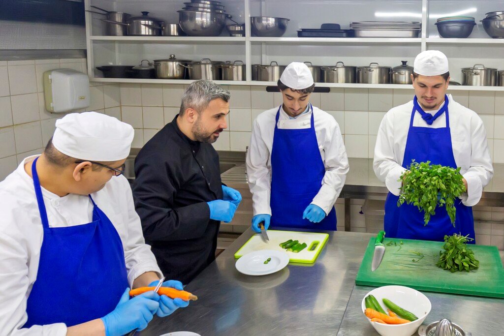 شيف يوجه ثلاثة طلاب بالزي الأبيض والمريول الأزرق في مطبخ تدريبي بمعهد الهادي | A chef instructing three students in white uniforms and blue aprons in a training kitchen at Al-Hadi Institute