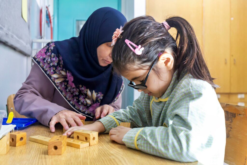 معلمة تساعد طفلة بنظارة وسماعة أذن على استخدام مكعبات تعليمية في مبرة الهادي | A teacher helping a girl with glasses and a hearing aid use educational blocks at Al-Hadi Mabarra
