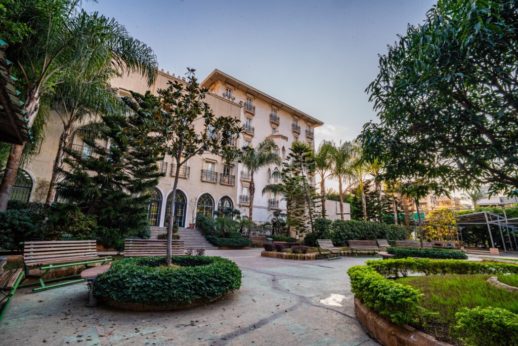 مبنى جامعي عريق تحيط به حديقة بأشجار النخيل ومقاعد خشبية | A stately university building surrounded by a garden with palm trees and wooden benches