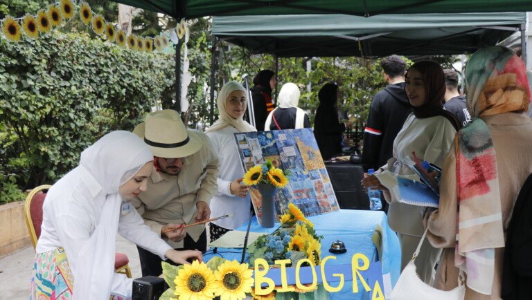 طلاب جامعة USAL حول طاولة مزينة بعباد الشمس ولوحات فنية في نشاط خارجي | USAL university students around a table decorated with sunflowers and artworks at an outdoor activity