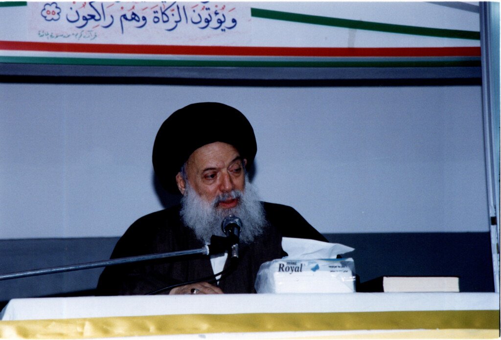 مؤسس جمعية المبرات يتحدث عبر الميكروفون وخلفه لافتة بآية قرآنية | The founder of Al-Mabarrat Association speaking into a microphone before a banner with a Quranic verse