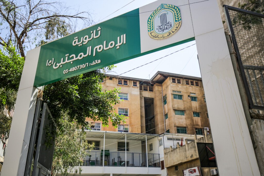 بوابة ثانوية الإمام المجتبى (ع) بلافتتها الخضراء وشعار جمعية المبرات | The gate of Al-Imam Al-Mujtaba High School with its green sign and Al-Mabarrat logo