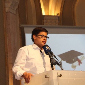 شاب بقميص أبيض يتحدث في الميكروفون خلف منبر زجاجي أمام شاشة تعرض قبعة تخرج | A young man in a white shirt speaks into the microphone behind a glass podium before a screen showing a graduation cap