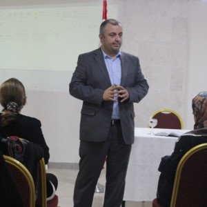 رجل ببدلة رمادية يقف متحدثاً أمام مشاركات محجبات جالسات في قاعة أمام شاشة عرض | A man in a grey suit stands addressing female participants in headscarves seated in a hall before a projection screen